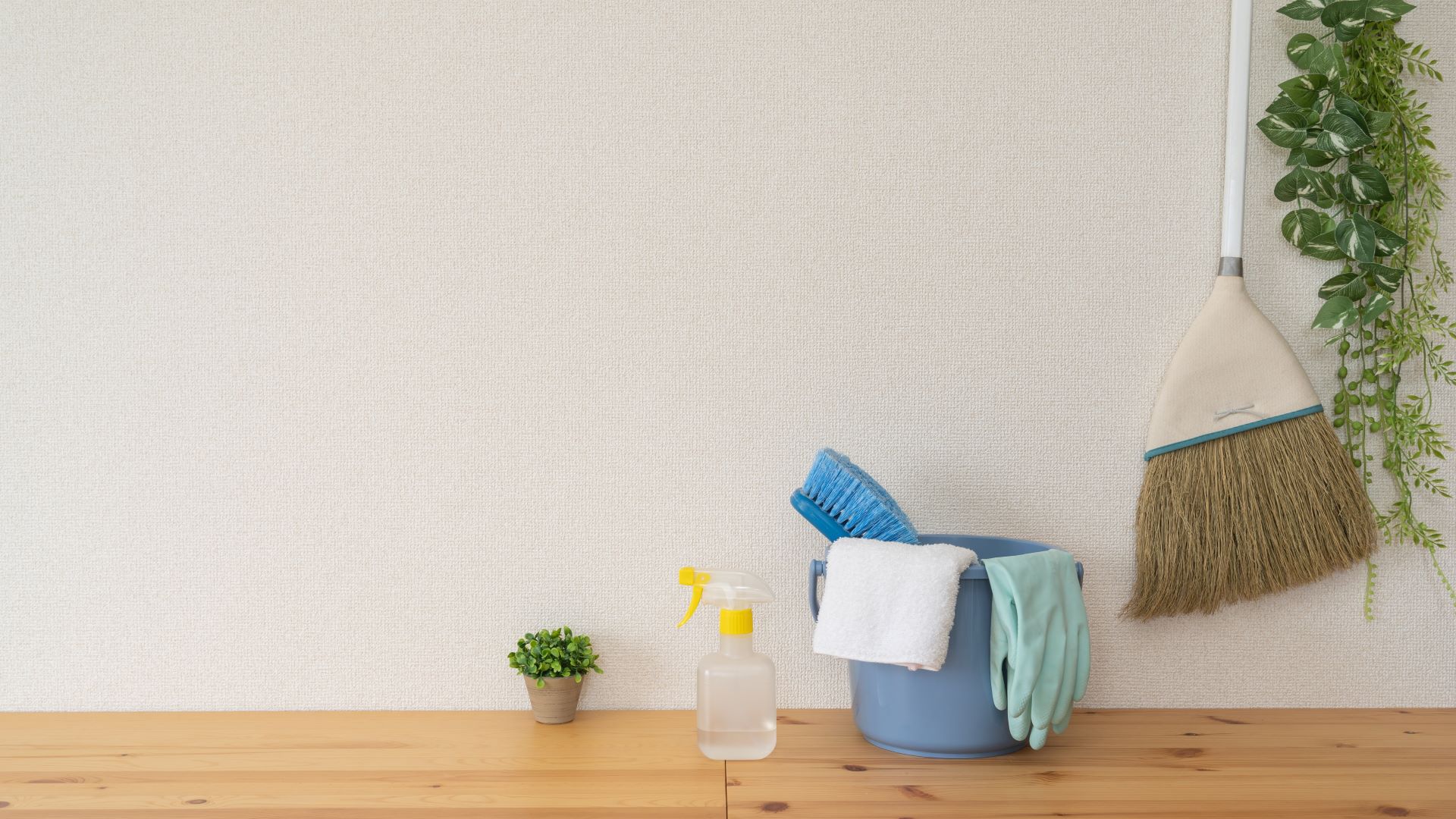 Ein Set von Reinigungswerkzeugen, darunter ein Eimer mit Bürste, Handschuhen und Sprühflasche, sowie ein Besen und eine Pflanze auf einem Holztisch, vor einer weißen Wand.
