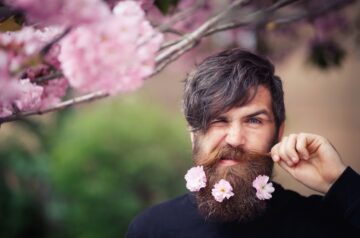 Mann mit dichtem Bart, geschmückt mit rosa Blüten, zwinkert in die Kamera vor einem blühenden Hintergrund