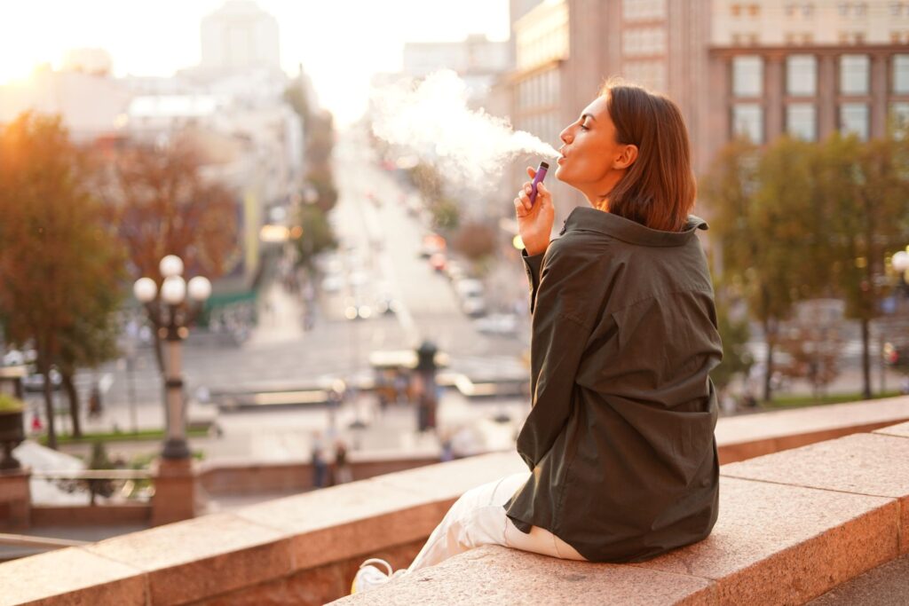 Junge Frau sitzt in der Stadt und genießt eine Einweg Vape mit Nikotin, zeigt entspannte Momente und moderne Freizeitgestaltung in urbaner Umgebung