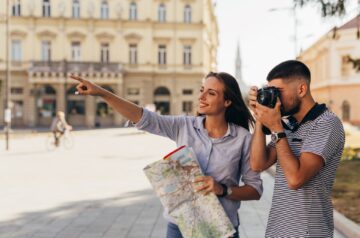 Ein Paar erkundet die Stadt, während die Frau eine Karte hält und auf ein Ziel zeigt, und der Mann mit einer Kamera ein Foto macht, bereit, besondere Momente festzuhalten.