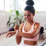 Junge Frau in Sportkleidung freut sich über Trainingserfolg und überprüft Fitnessdaten auf Smartwatch, modernes Fitnessstudio im Hintergrund