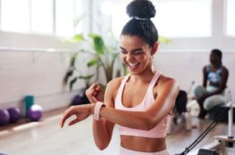 Junge Frau in Sportkleidung freut sich über Trainingserfolg und überprüft Fitnessdaten auf Smartwatch, modernes Fitnessstudio im Hintergrund