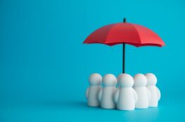 Weisse Spielfiguren stehen unter rotem Schirm – abstraktes Symbol fuer Schutz, Betreuung und Familienservice im Unternehmen