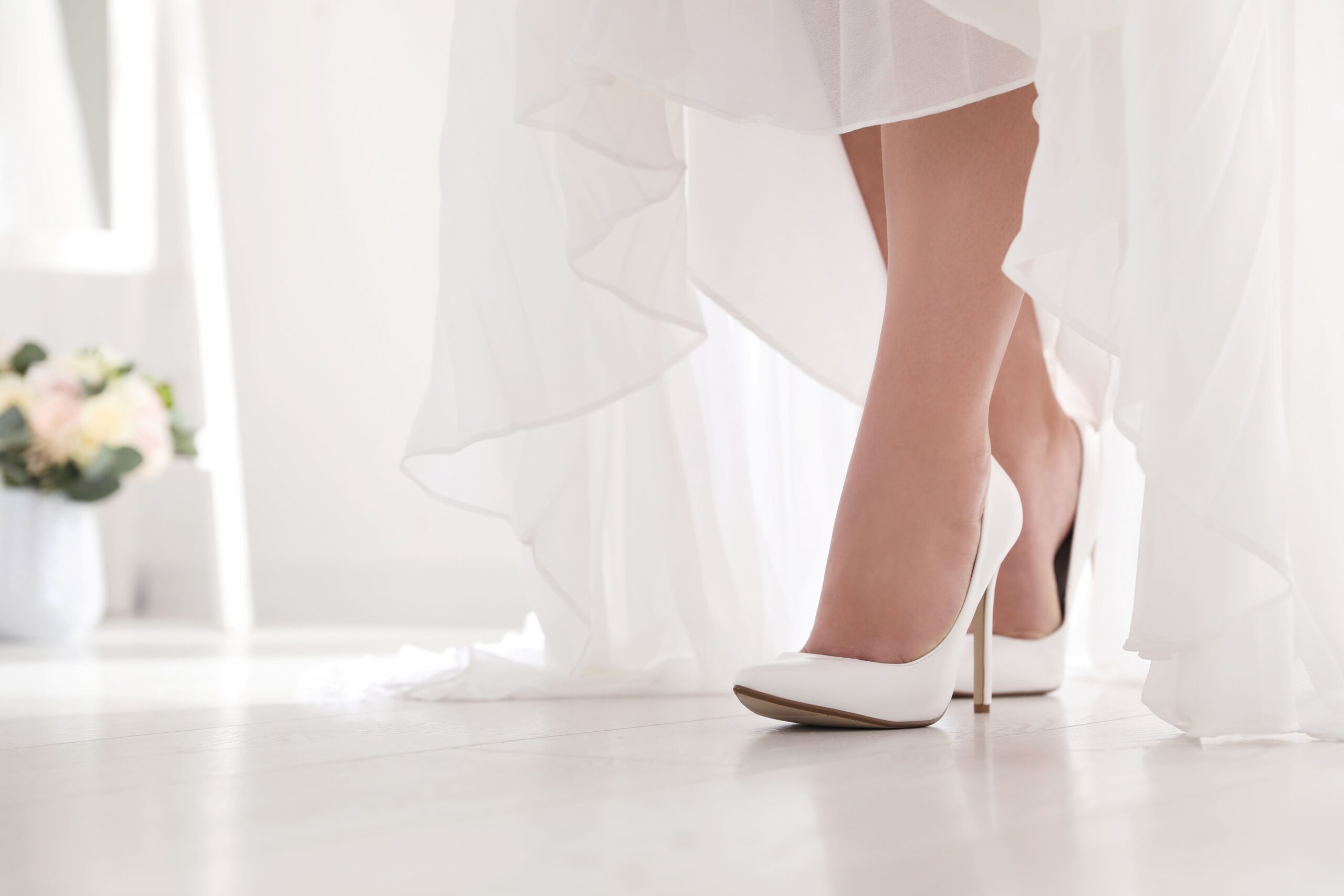 Brautschuhe beim Anprobieren – Braut steht in weißen High Heels auf hellem Boden im Hochzeitskleid