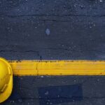 Gelber Schutzhelm liegt auf nasser gelber Linie einer Bodenmarkierung auf Asphalt – Symbol fuer Sicherheit und Struktur