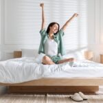 Frau sitzt morgens entspannt auf einem großen Bett mit weißem Spannbettlaken 200x200 in hellem, minimalistisch eingerichteten Schlafzimmer