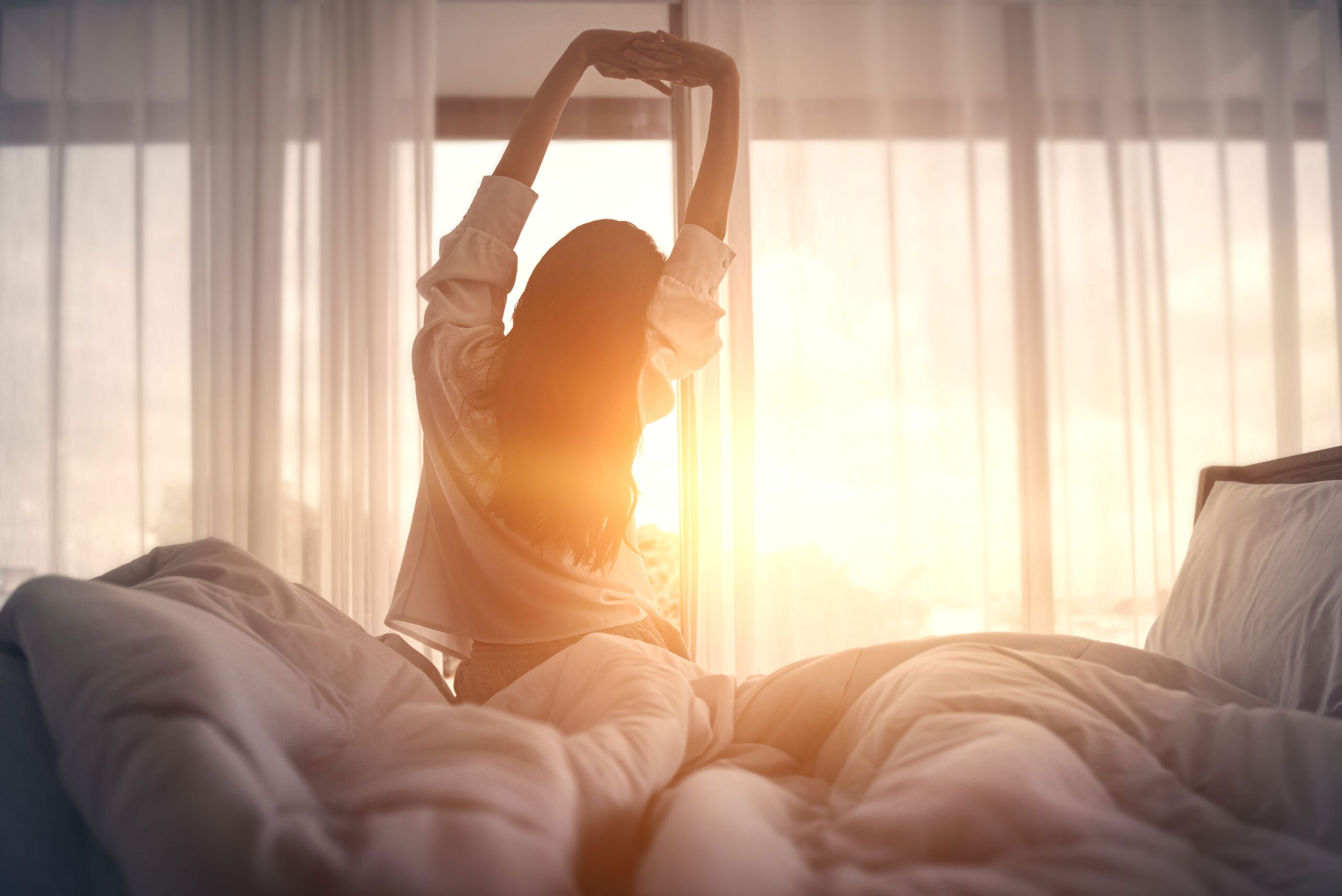 Frau sitzt im Morgenlicht auf einem Bett mit Spannbettlaken, umgeben von weichen Textilien und entspannter Schlafzimmeratmosphäre